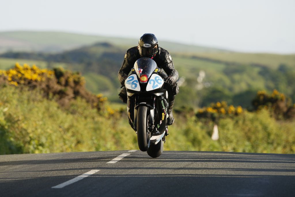 A motorcyclist performs a wheelie on a scenic road in the Isle of Man during the day.