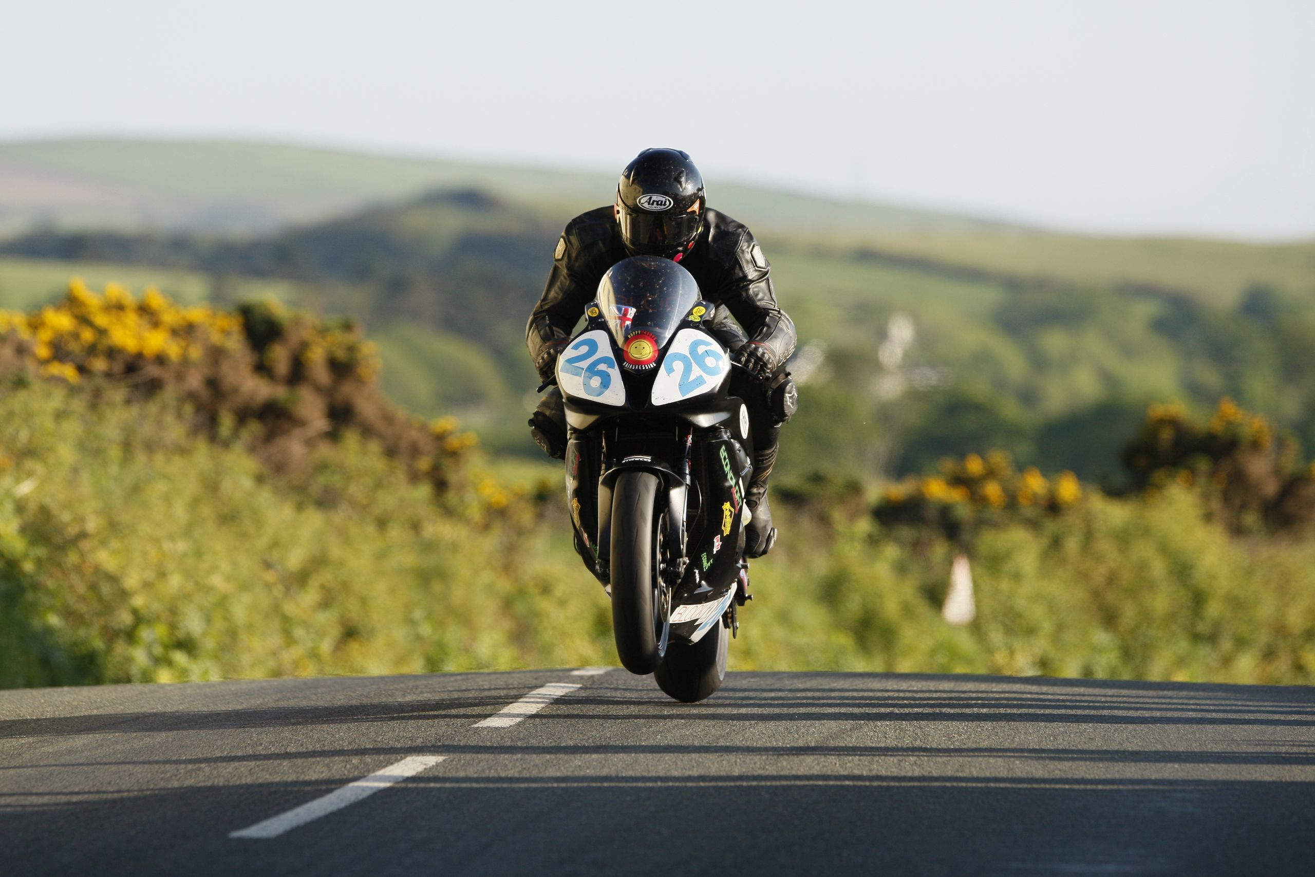 A motorcyclist performs a wheelie on a scenic road in the Isle of Man during the day.