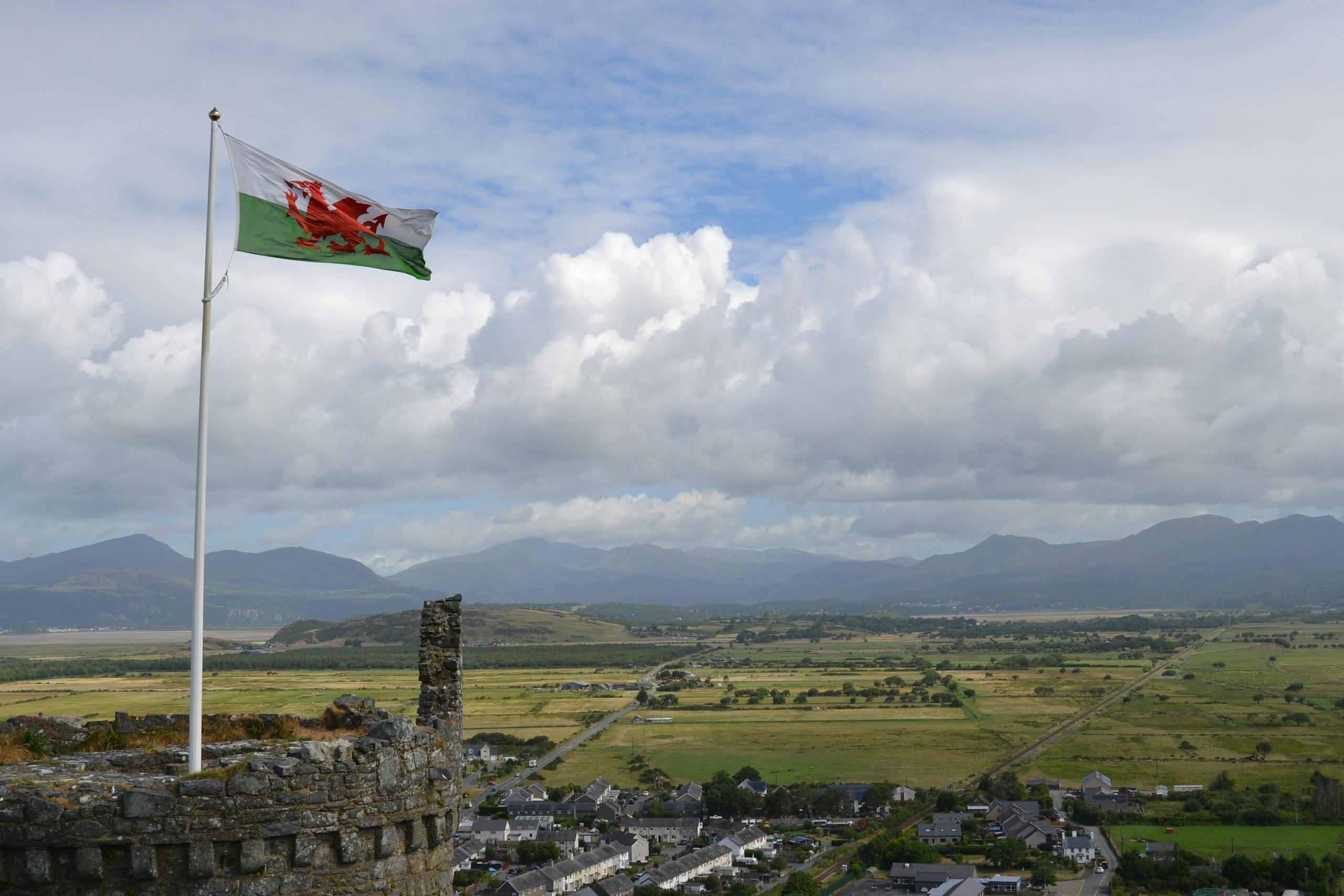 A Welsh flag waves over a scenic countryside landscape with mountains and a town visible.