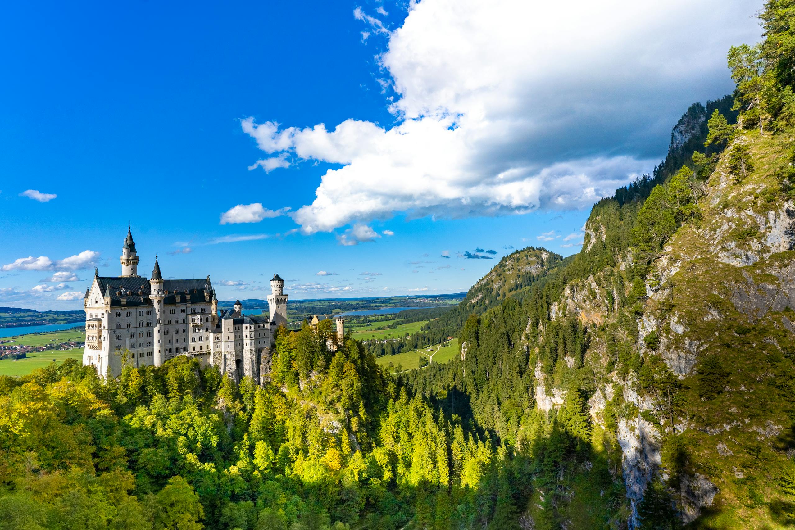 Aerial view of Neuschwanstein Castle in Bavaria, Germany, surrounded by lush forests and mountains under a blue sky.