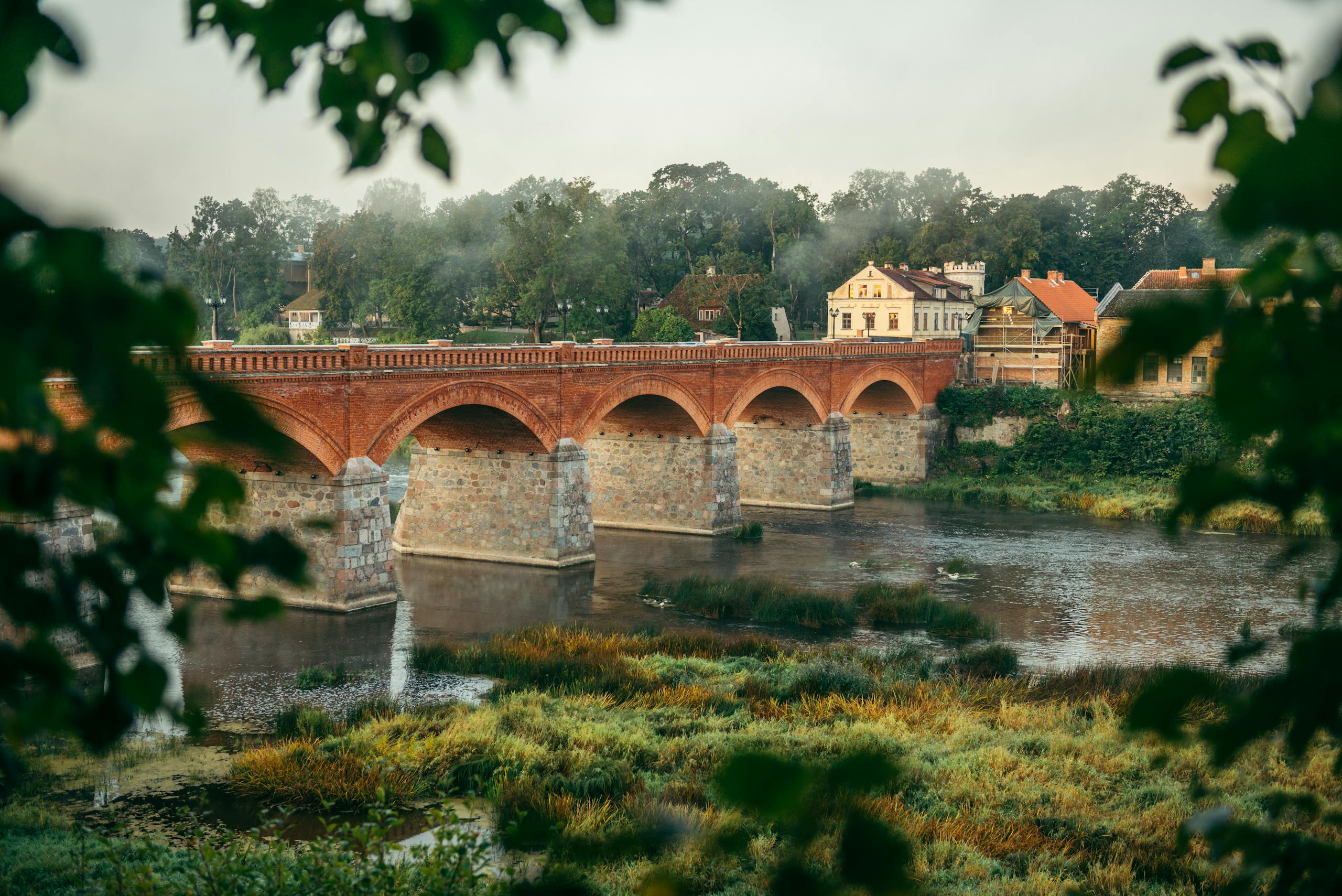 Historic red brick bridge over river in Kuldīga, Latvia surrounded by lush greenery.